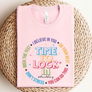 May include: A light pink t-shirt with a round neckline. The shirt features a colorful graphic with the words "TIME TO LOCK IN" in the center, surrounded by phrases like "I BELIEVE IN YOU" and "YOU CAN DO THIS". The hashtag "#testday" is also included.
