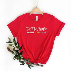 May include: A red t-shirt with the text "We The People MEANS EVERYONE" in white and rainbow colors. The shirt is displayed on a wooden hanger against a white background, with a sprig of greenery at the bottom.