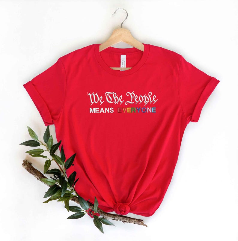May include: A red t-shirt with the text "We The People MEANS EVERYONE" in white and rainbow colors. The shirt is displayed on a wooden hanger against a white background, with a sprig of greenery at the bottom.