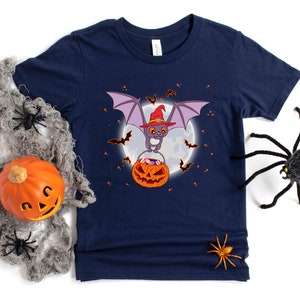 May include: A navy blue t-shirt with a cartoon bat wearing a witch hat and holding a pumpkin full of candy. The bat is flying in front of a full moon with stars in the background.