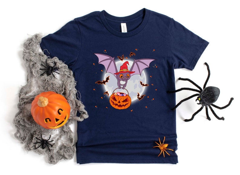 May include: A navy blue t-shirt with a cartoon bat wearing a witch hat and holding a pumpkin full of candy. The bat is flying in front of a full moon with stars in the background.