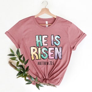 May include: A mauve t-shirt with a colorful graphic that reads "He is Risen" with the scripture reference "Matthew 28:6" underneath.