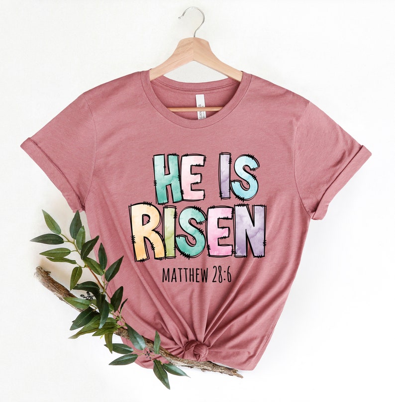 May include: A mauve t-shirt with a colorful graphic that reads "He is Risen" with the scripture reference "Matthew 28:6" underneath.