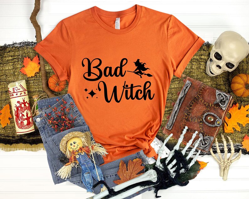 Puede incluir: Camiseta naranja con el texto "Bad Witch" en letras cursivas negras. Una silueta de bruja vuela en una escoba sobre el texto. La camiseta est&aacute; rodeada de decoraciones de Halloween, incluyendo una calavera, un libro y hojas de oto&ntilde;o.