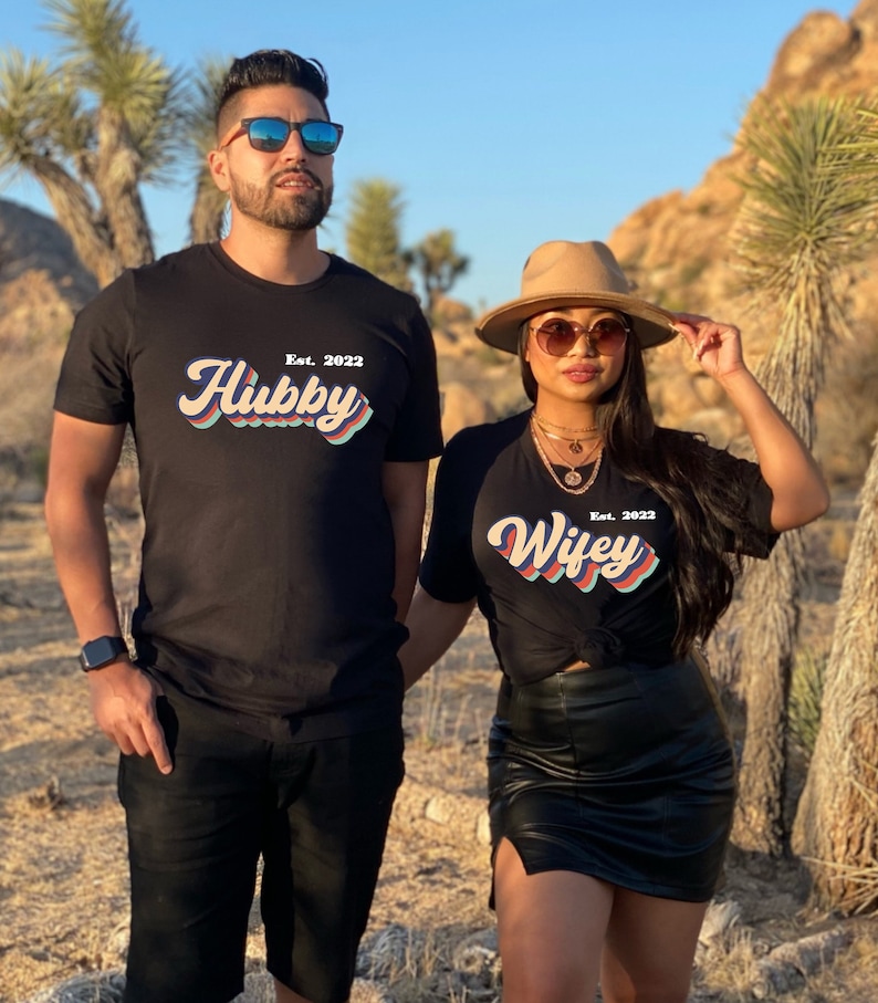 May include: A couple wearing matching black t-shirts with the text "Hubby Est. 2022" and "Wifey Est. 2022" in a retro font style. The shirts are a dark black color and the text is in a multi-colored rainbow design.