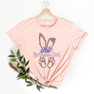 K&ouml;nnte beinhalten: Ein pinkfarbenes T-Shirt mit einem Aufdruck von Hasenohren und -pfoten mit Leopardenmuster. Die Hasenohren haben ein Blumenband mit lila Blumen. Der Text "Birthday Girl" ist auf das T-Shirt gedruckt.