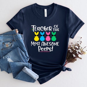May include: Navy blue t-shirt with the text "Teacher of the Most Awesome Peeps!" and colorful bunny graphics wearing sunglasses. The shirt is paired with blue jeans, suggesting a casual, everyday style. The design is perfect for Easter or a teacher appreciation gift.