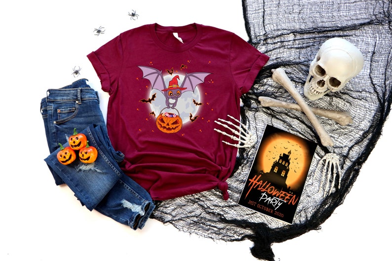 May include: A maroon t-shirt with a graphic of a bat wearing a witch's hat and holding a jack-o'-lantern. The bat is flying in front of a full moon with bats flying around it. The t-shirt is laid out on a white background with a pair of blue jeans, a skull, and a Halloween party invitation.