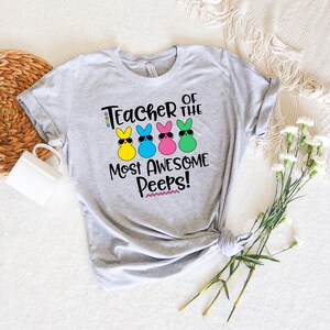 May include: A light gray t-shirt with the text "Teacher of the Most Awesome Peeps!" and four colorful bunny graphics wearing sunglasses. The bunnies are yellow, blue, pink, and green.