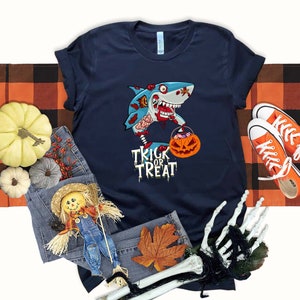 May include: A navy blue t-shirt with a graphic of a zombie shark holding a pumpkin bucket. The text "Trick or Treat" is printed below the shark.