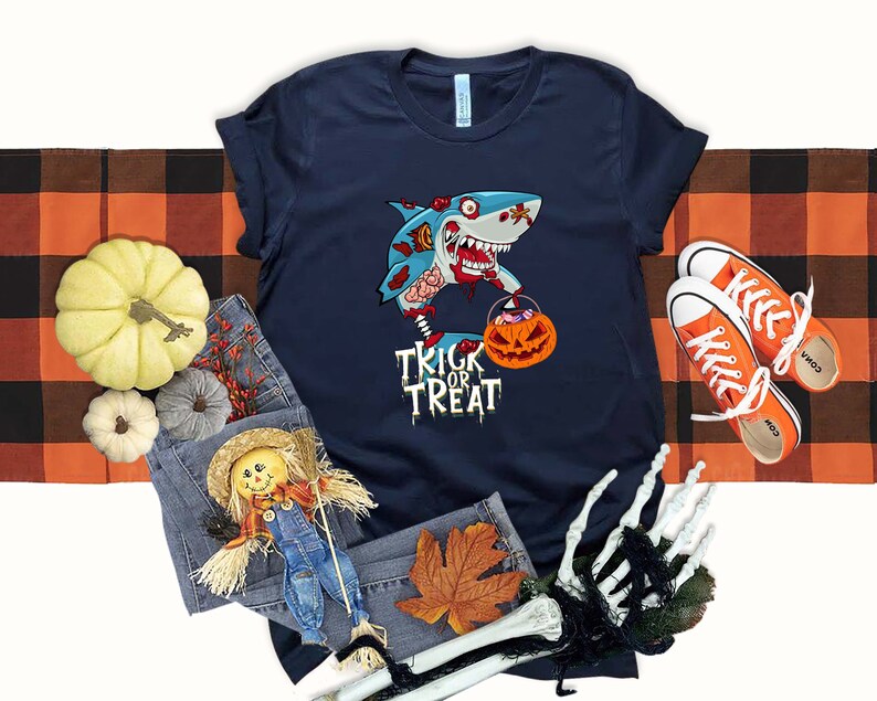 May include: A navy blue t-shirt with a graphic of a zombie shark holding a pumpkin bucket. The text "Trick or Treat" is printed below the shark.