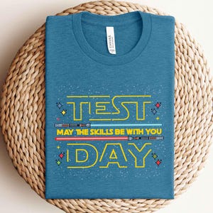 May include: Teal t-shirt with the words "TEST DAY" in yellow, along with the phrase "MAY THE SKILLS BE WITH YOU." The design includes blue and red lightsabers. The shirt is displayed on a woven, circular surface.