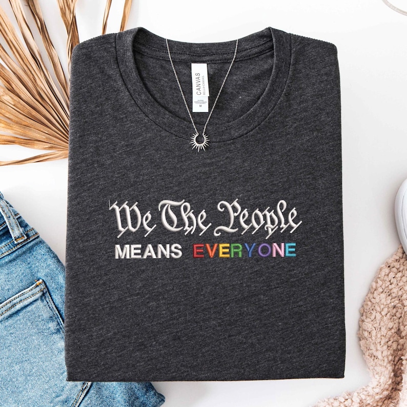 May include: Dark gray t-shirt with the embroidered text "We The People MEANS EVERYONE" in white and rainbow colors. A silver necklace with a sun pendant is on the shirt. The shirt is folded on a white surface.