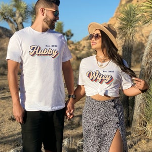 May include: A couple wearing white t-shirts with the text "Hubby Est. 2022" and "Wifey Est. 2022" in a retro font style. The text is in a rainbow color scheme. The couple is holding hands and smiling at each other.