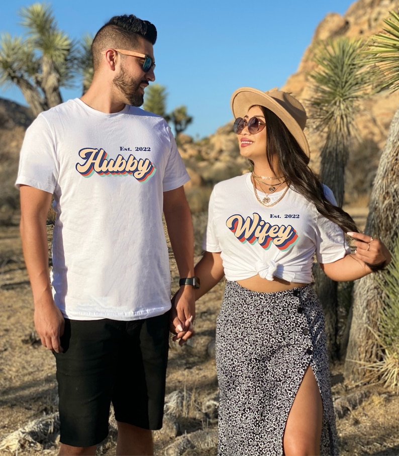 May include: A couple wearing white t-shirts with the text "Hubby Est. 2022" and "Wifey Est. 2022" in a retro font style. The text is in a rainbow color scheme. The couple is holding hands and smiling at each other.
