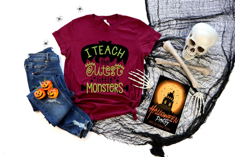 May include: A maroon t-shirt with the text "I teach the cutest little monsters" in black and green lettering. The shirt is laid out on a white surface with a pair of blue jeans, three orange pumpkins, and a black and white Halloween party invitation.