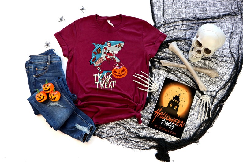 May include: A maroon t-shirt with a graphic of a zombie shark holding a pumpkin bucket with the text "Trick or Treat". The shirt is laid out on a white surface with a pair of blue jeans, a skull, and a Halloween party invitation.