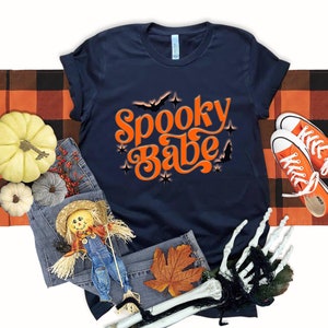 May include: A navy blue t-shirt with the text "Spooky Babe" in orange with a retro font. The text is surrounded by stars and bats. The shirt is laid out on a black and orange checkered fabric.