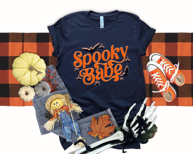 May include: A navy blue t-shirt with the text "Spooky Babe" in orange with a retro font. The text is surrounded by stars and bats. The shirt is laid out on a black and orange checkered fabric.