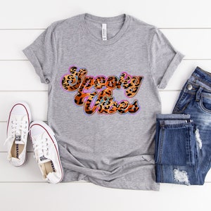 May include: A gray t-shirt with the text "Spooky Vibes" in a leopard print font with a pink, blue, and orange outline. The shirt is laying on a white wooden surface with a pair of white sneakers and blue jeans.