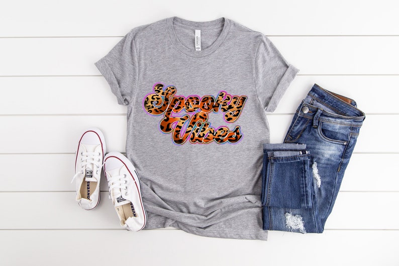 May include: A gray t-shirt with the text "Spooky Vibes" in a leopard print font with a pink, blue, and orange outline. The shirt is laying on a white wooden surface with a pair of white sneakers and blue jeans.