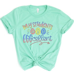 May include: A light green t-shirt with the text "My Students are Eggcellent" in rainbow colors. The text includes three stylized eggs with the letters "A", "R", and "E" inside.
