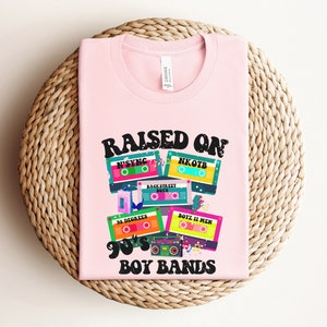 May include: Pink t-shirt with the text "RAISED ON 90'S BOY BANDS" and a graphic of cassette tapes with the names of boy bands from the 1990s, including NSYNC, NKOTB, Backstreet Boys, 98 Degrees, and Boyz II Men.