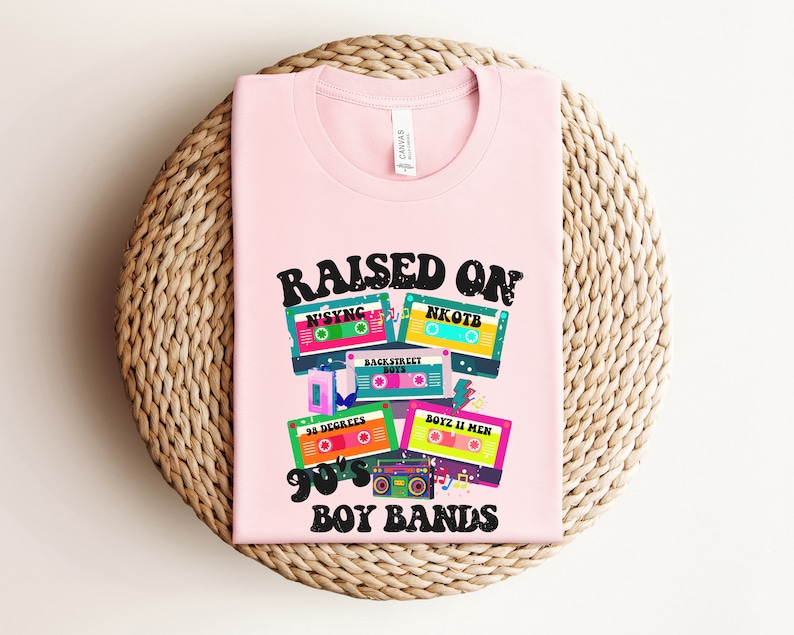 May include: Pink t-shirt with the text "RAISED ON 90'S BOY BANDS" and a graphic of cassette tapes with the names of boy bands from the 1990s, including NSYNC, NKOTB, Backstreet Boys, 98 Degrees, and Boyz II Men.