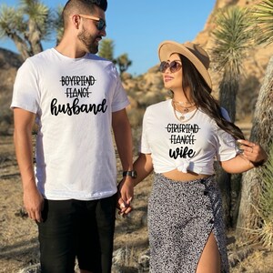 May include: A couple wearing white t-shirts with the text "Boyfriend Fiance Husband" and "Girlfriend Fiance Wife" printed on them. They are holding hands and smiling at each other.