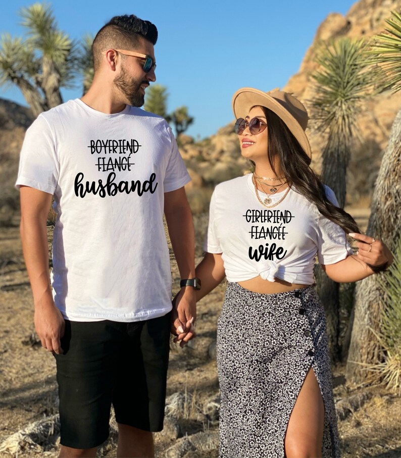 May include: A couple wearing white t-shirts with the text "Boyfriend Fiance Husband" and "Girlfriend Fiance Wife" printed on them. They are holding hands and smiling at each other.