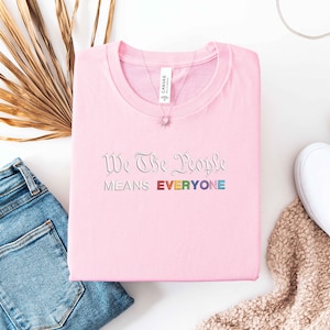 May include: A light pink t-shirt with the embroidered text "We The People Means EVERYONE" in a rainbow color scheme. The shirt is folded neatly, with a necklace and white sneakers visible.