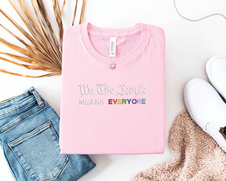 May include: A light pink t-shirt with the embroidered text "We The People Means EVERYONE" in a rainbow color scheme. The shirt is folded neatly, with a necklace and white sneakers visible.