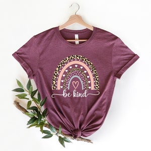 Op de afbeelding: Een maroon t-shirt met een regenboogontwerp met een luipaardprint, roze en witte hartjes en de tekst "be kind".