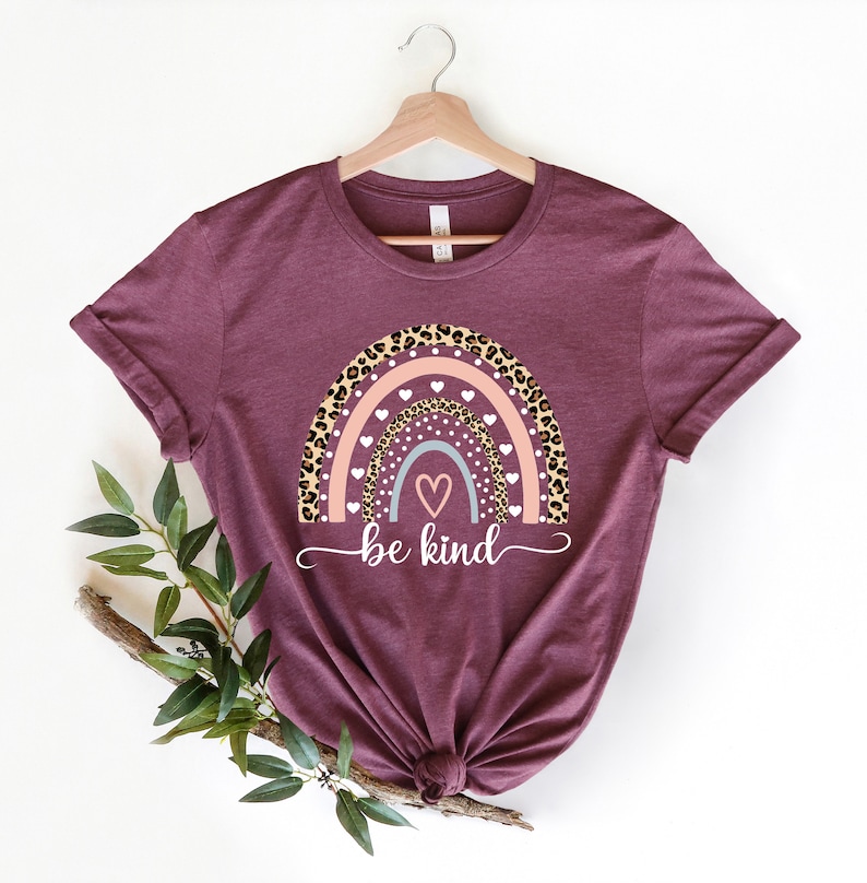 Op de afbeelding: Een maroon t-shirt met een regenboogontwerp met een luipaardprint, roze en witte hartjes en de tekst "be kind".