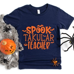 May include: A navy blue t-shirt with the text "SPOOK TAKULAR TEACHER" in orange. The shirt is decorated with bats and spiders.