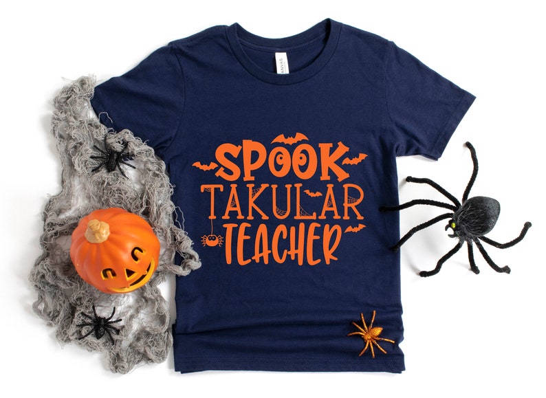 May include: A navy blue t-shirt with the text "SPOOK TAKULAR TEACHER" in orange. The shirt is decorated with bats and spiders.
