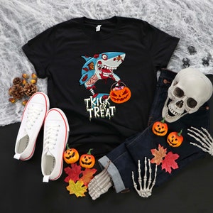 May include: Black t-shirt with a cartoon zombie shark holding a pumpkin bucket and the text "Trick or Treat". The shirt is laid out on a black surface with a pair of white sneakers, a skull, and a skeleton hand.