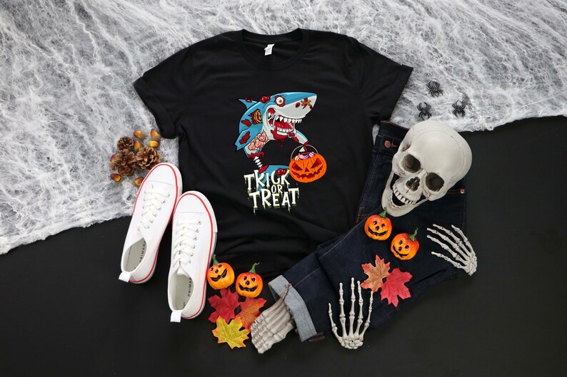 May include: Black t-shirt with a cartoon zombie shark holding a pumpkin bucket and the text "Trick or Treat". The shirt is laid out on a black surface with a pair of white sneakers, a skull, and a skeleton hand.