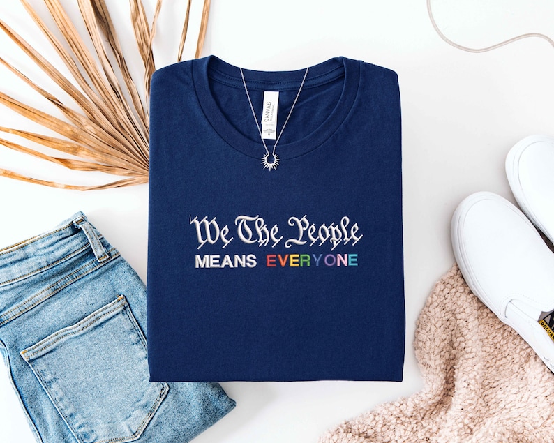 May include: Navy blue t-shirt with the text "We The People MEANS EVERYONE" in white and rainbow colors. A silver necklace with a sunburst pendant rests on the shirt. The shirt is displayed with blue jeans and white slip-on shoes.