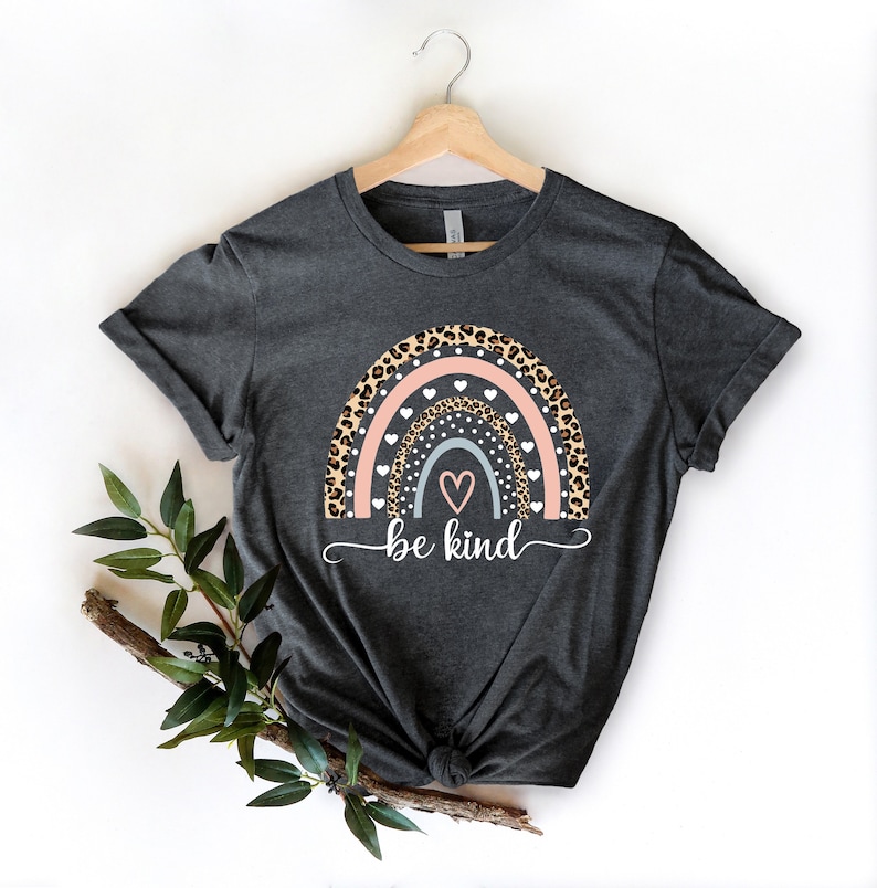 Op de afbeelding: Een donkergrijs T-shirt met een regenboogontwerp met een luipaardprint, witte stippen en roze en blauwe harten. De tekst "be kind" is onder de regenboog gedrukt.