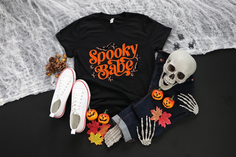May include: Black t-shirt with the text "Spooky Babe" in orange lettering. The shirt is surrounded by Halloween decorations, including a skull, pumpkins, and fall leaves.