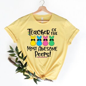 May include: Yellow t-shirt with the text "Teacher of the Most Awesome Peeps!" and four cartoon bunny figures wearing sunglasses. The bunnies are yellow, blue, pink, and green. The shirt is hanging on a wooden hanger.