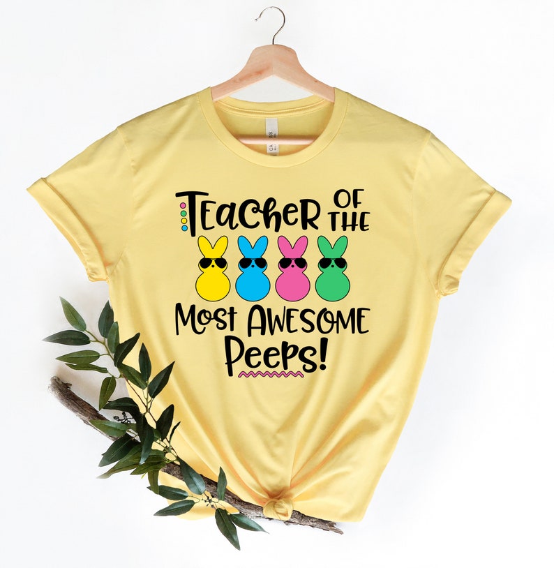 May include: Yellow t-shirt with the text "Teacher of the Most Awesome Peeps!" and four cartoon bunny figures wearing sunglasses. The bunnies are yellow, blue, pink, and green. The shirt is hanging on a wooden hanger.
