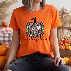 Puede incluir: Camiseta naranja con un gr&aacute;fico de una calabaza con un coraz&oacute;n blanco y el texto "hello fall".