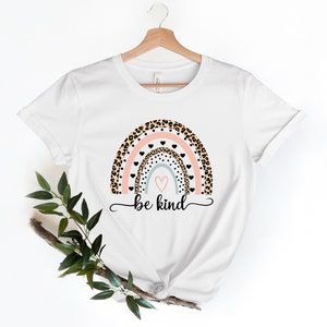 Op de afbeelding: Wit T-shirt met een regenboog-print met luipaardprint, roze en blauwe strepen en zwarte hartjes. De tekst "be kind" is onder de regenboog gedrukt.