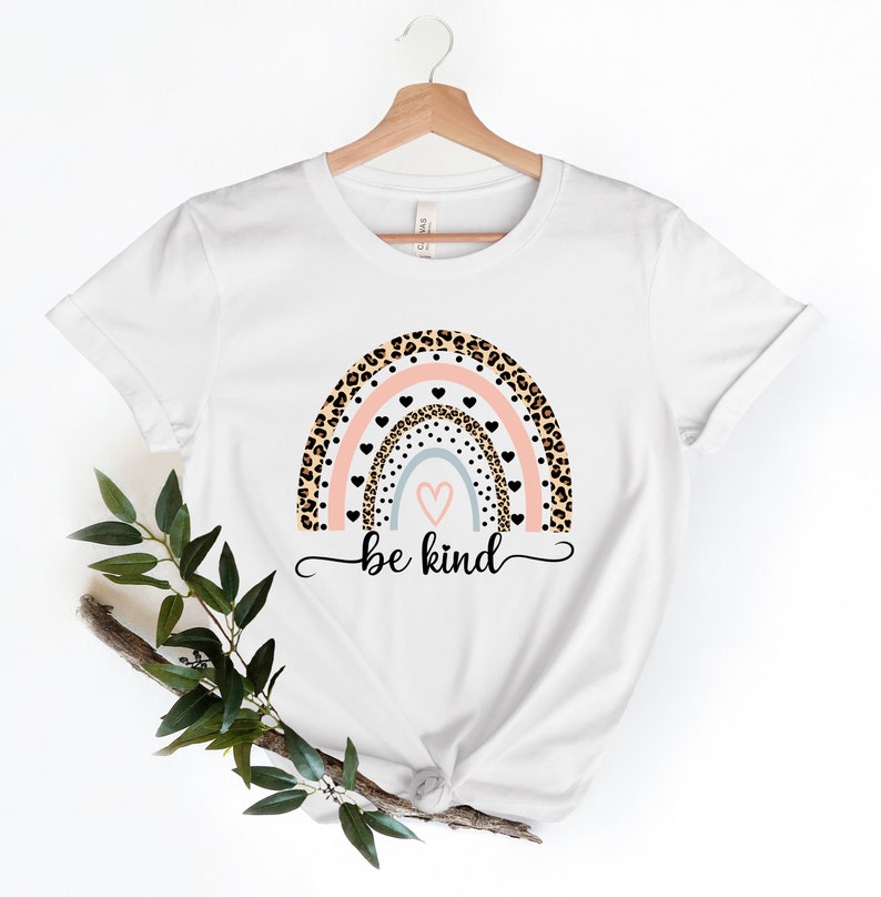 Op de afbeelding: Wit T-shirt met een regenboog-print met luipaardprint, roze en blauwe strepen en zwarte hartjes. De tekst "be kind" is onder de regenboog gedrukt.