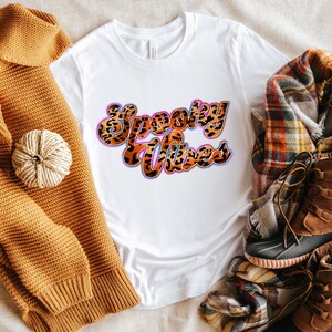 May include: White t-shirt with the text "Spooky Vibes" in a retro font with a leopard print pattern. The text is in pink, blue, and orange with a white outline.