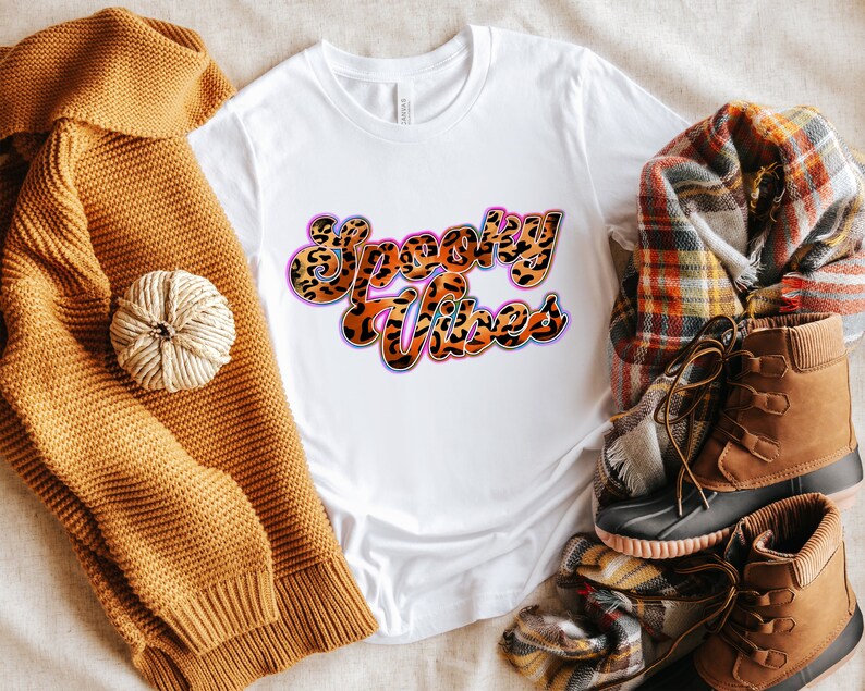 May include: White t-shirt with the text "Spooky Vibes" in a retro font with a leopard print pattern. The text is in pink, blue, and orange with a white outline.