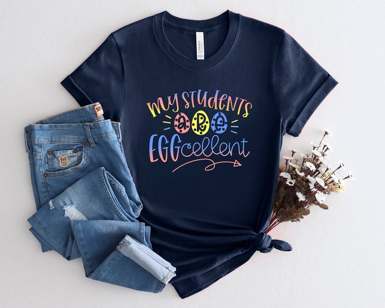 May include: A navy blue t-shirt with the text "My Students Are Eggcellent" in rainbow colors. The text is surrounded by three colorful eggs with white polka dots.