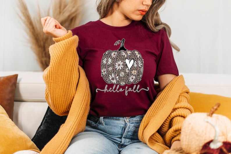 Puede incluir: Una camiseta color burdeos con un dise&ntilde;o de calabaza floral blanco y negro y el texto "hello fall".